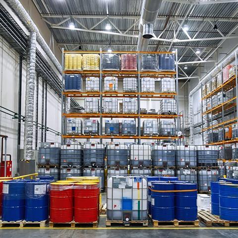Oil drums and plastic container on pallets in a warehouse on metal shelving.